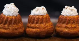 Three small golden cupcakes with white whipped cream frosting and tiny pearl sprinkles lined up on a dark surface
