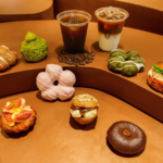 Assorted donuts and pastries arranged on curved brown steps with two coffee drinks in the background cafe display