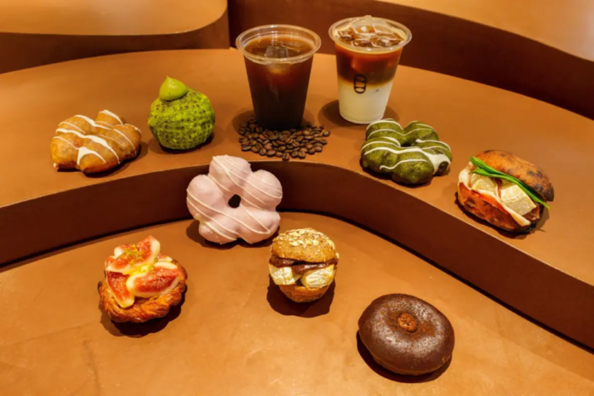 Assorted donuts and pastries arranged on curved brown steps with two coffee drinks in the background cafe display