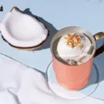 Copper mug with whipped coconut cream topped with toasted coconut on a pastel blue surface beside a halved coconut