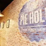 Brick walled hallway with industrial pendant lights and framed pictures on the left a large blue mural with the text the HOLE on the right wall