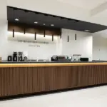 Modern coffee counter with a wooden slat front black countertop and hanging pendant lights in a bright minimalist lobby Sign reads Shikishima Coffee Counter