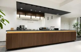 Modern coffee counter with a wooden slat front black countertop and hanging pendant lights in a bright minimalist lobby Sign reads Shikishima Coffee Counter