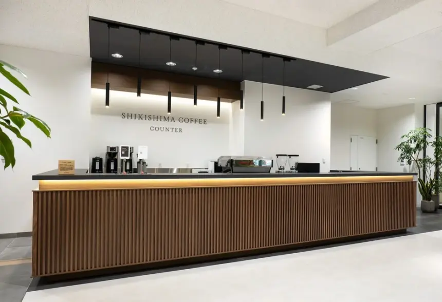 Modern coffee counter with a wooden slat front black countertop and hanging pendant lights in a bright minimalist lobby Sign reads Shikishima Coffee Counter