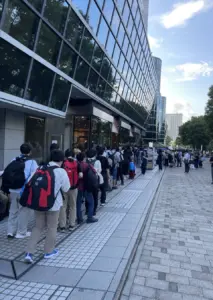 People with backpacks line up along the sidewalk outside a glass fronted building on a sunny day