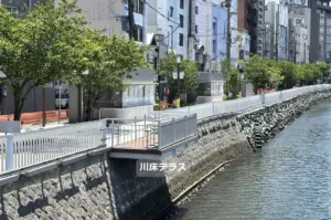 Riverside promenade with trees white railing and a wooden terrace over a stone embankment beside an urban canal with buildings in the background in Japanese text 川床テラス on the terrace edge