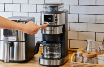 Hand pressing a button on a stainless steel coffee grinder beside a drip coffee maker in a kitchen