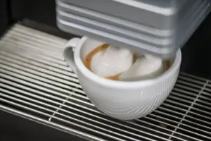 White ceramic cup under an espresso machine with two mounds of milk foam floating on a small coffee drink on a metal grate