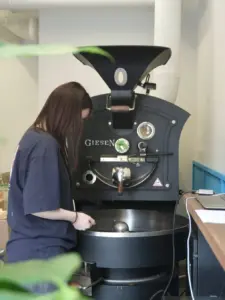 A woman in a navy uniform operates a large Giesen coffee roaster beans cooling in a tray beneath the machine in a processing room