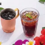 Iced coffee in a copper mug and a glass of iced tea on a white table surrounded by hibiscus and plumeria flowers with a cafe logo in the top left