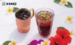Iced coffee in a copper mug and a glass of iced tea on a white table surrounded by hibiscus and plumeria flowers with a cafe logo in the top left