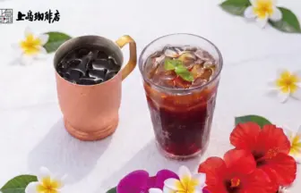 Iced coffee in a copper mug and a glass of iced tea on a white table, surrounded by hibiscus and plumeria flowers, with a cafe logo in the top-left.