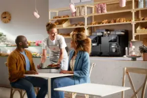 A smiling server in an apron hands a tray with coffee and food to a couple seated at a cafe table