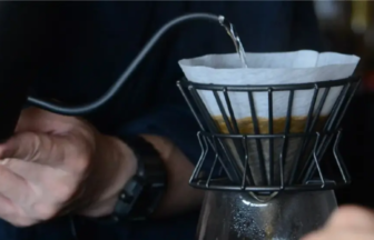 Close-up of hands pouring hot water through a paper-filtered metal dripper into a glass carafe for pour-over coffee.