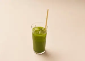 Tall glass of bright green juice with ice and a wooden straw on a pale beige background