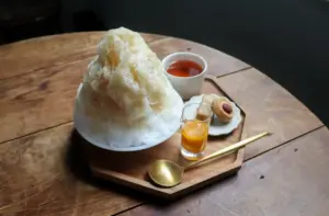 Cone shaped shaved ice dessert topped with syrup on a wooden board accompanied by tea orange juice a cookie and a small spoon