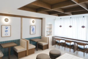 Bright cafe interior with booth seating along a teal tiled wall and a long bar counter with wooden stools by a large window with sheer curtains