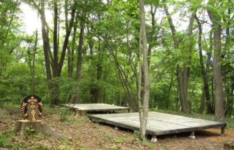 Wooden stage platforms in a green forest; a circular sign resembling the Starbucks logo sits on a tree stump.