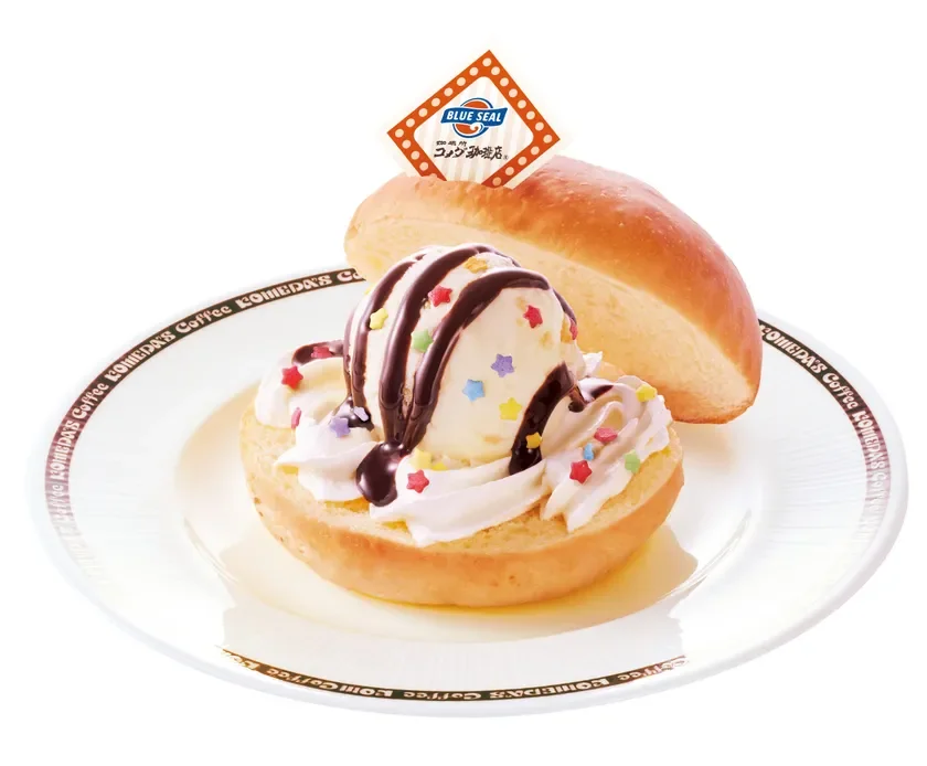 白い皿に盛られたデザートで、丸いパンの上にバニラアイスとホイップクリーム、チョコレートソースとカラフルな星形スプリンクルがトッピングされています。奥にはもう一つのパンが添えられ、ブルーシールのロゴが入った旗が立っています。コメダ珈琲店のロゴが入った皿を使用しています。