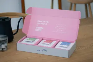 Pink open box with three sachets in separate compartments on a wooden table a black kettle sits to the left
