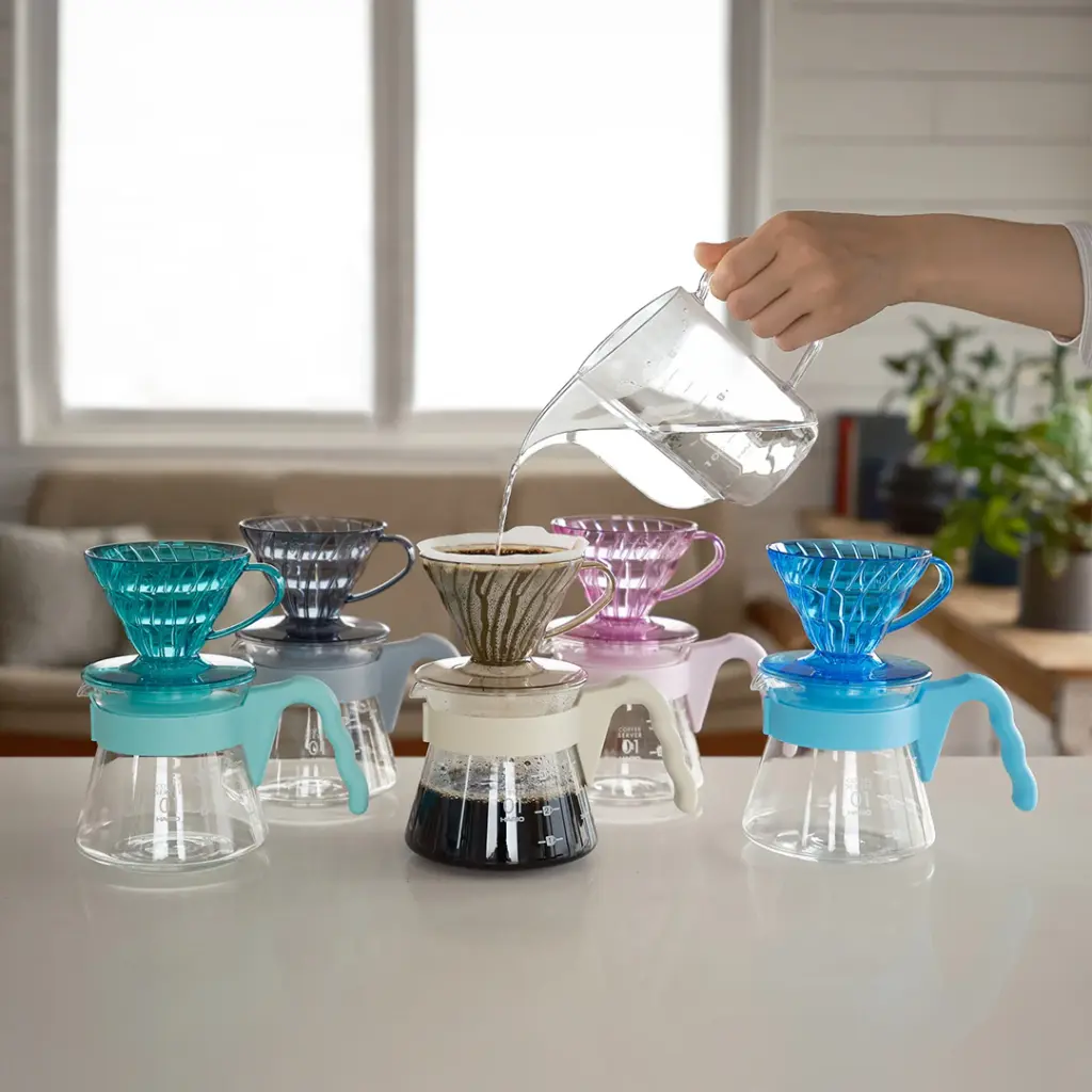 Person pours hot water into a pour over coffee setup with six glass carafes and colorful drippers on a white countertop in a bright kitchen