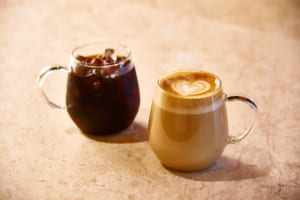 Two glass mugs on a light marble surface a dark iced coffee on the left and a creamy latte with a heart shaped foam design on the right