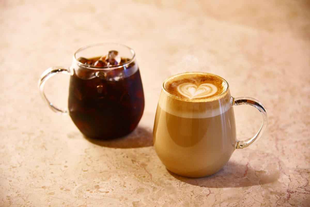 Two glass mugs on a light marble surface a dark iced coffee on the left and a creamy latte with a heart shaped foam design on the right