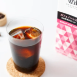 Glass of iced coffee with ice cubes on a cork coaster next to a pink decaf iced coffee carton on a white surface
