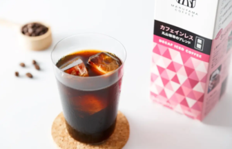 Glass of iced coffee with ice cubes on a cork coaster next to a pink decaf iced coffee carton on a white surface