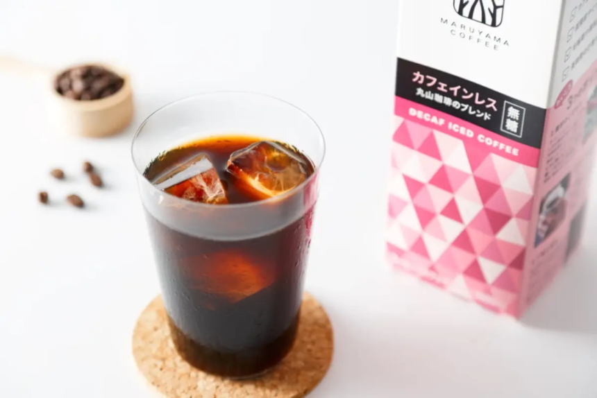 Glass of iced coffee with ice cubes on a cork coaster next to a pink decaf iced coffee carton on a white surface