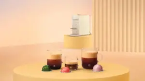 White coffee machine on a yellow round pedestal with three glass mugs of coffee and colorful coffee capsules on a pastel set