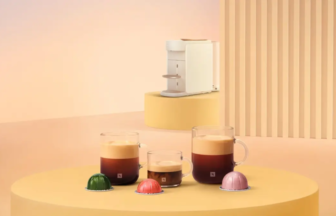 White coffee machine on a yellow circular platform surrounded by three glass mugs of espresso and three pastel coffee capsules on a pale yellow table