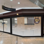 Cafe de CRIÉ storefront in a mall with dark brown framing frosted glass walls and a round logo seating is visible inside