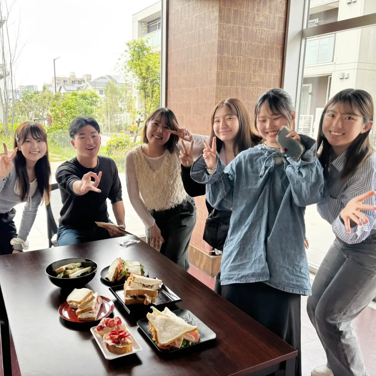 明るい室内で、6人の友人グループが笑顔で集まり、テーブルに並べられたサンドイッチや軽食を楽しんでいる様子