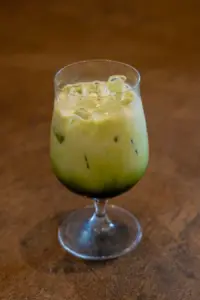 Green iced cocktail in a stemmed glass with ice on a brown tabletop likely a matcha or kiwi drink
