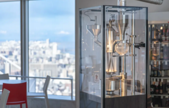 Glass enclosed coffee roaster in a bright cafe with a city skyline visible through large windows behind it