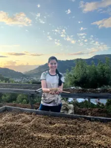 Smiling woman holding a handful of drying beans on a raised bed with hills and a sunset sky behind her on a farm path