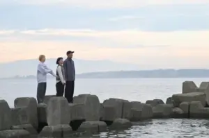 Three people standing on large concrete breakwaters along a calm sea at sunset looking toward the horizon