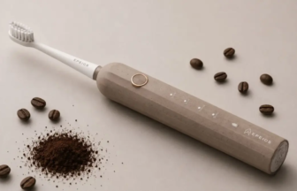 Beige electric toothbrush lying diagonally on a light surface with scattered coffee beans and a mound of ground coffee nearby and a ring on the body