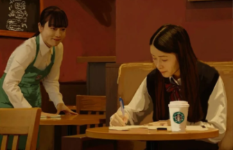 Two young women in a cozy cafe a student in a plaid skirt sits at a round table writing in a notebook with a Starbucks cup nearby while a waiter in a green apron stands by the table