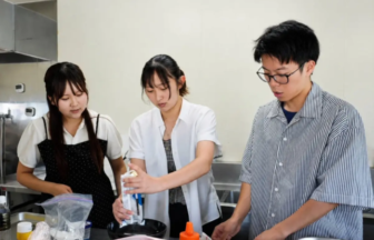 Three people in a bright kitchen: one pours liquid into a bowl as two others watch.