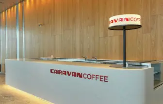 White marble lit Caravan Coffee counter with red logo modern wooden paneled wall and a circular lighted sign on a tall black pole behind the counter