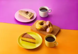 Colorful table with cake slice on a pink plate a cream filled pastry on a wooden board a cheesecake slice on a yellow plate and two cups of black coffee