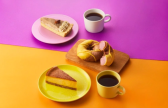 Slice of cheesecake on a pink plate, a glazed donut on a wooden board, and two cups of black coffee on a pink-orange backdrop with a yellow plate nearby on the table surface