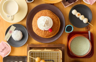 Top down view of a wooden table filled with desserts a pancake with cream center fruit bowl with kiwi and bananas matcha drinks dango skewers and assorted sweets on colorful plates and trays