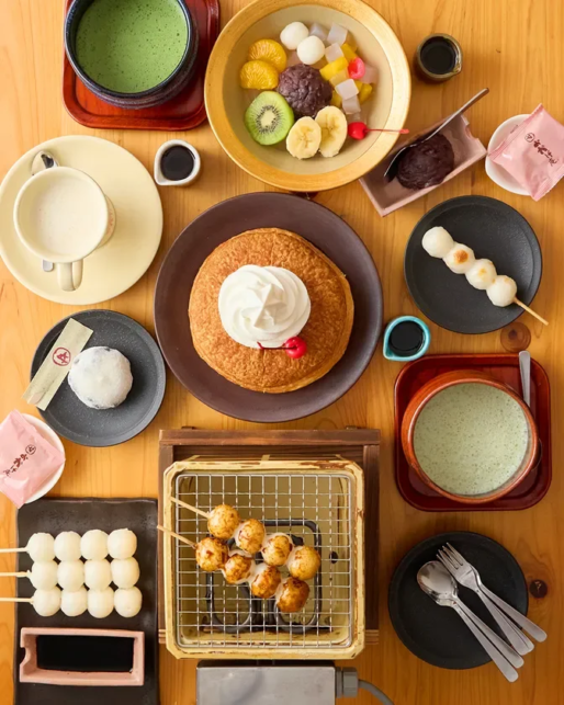Top down view of a wooden table filled with desserts a pancake with cream center fruit bowl with kiwi and bananas matcha drinks dango skewers and assorted sweets on colorful plates and trays
