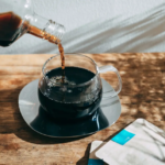 Dark coffee is being poured from a bottle into a clear glass mug on a wooden table with sugar packets nearby on the surface