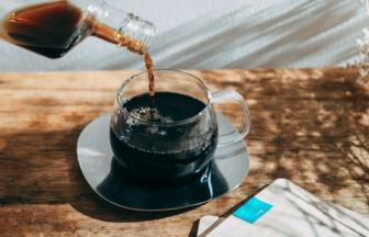 Dark coffee is being poured from a bottle into a clear glass mug on a wooden table, with sugar packets nearby on the surface.
