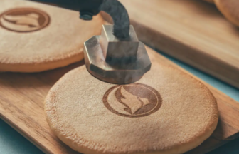 Close-up of a metal stamp pressing into a round wooden disc with a circular flame logo being imprinted.