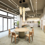 Open plan loungedining area with a round table and wooden chairs large glass doors and potted plants across a concrete ceiling space soft lighting